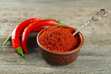 Photo of Bowl of ground red pepper with spoon and ingredient on wooden table, closeup