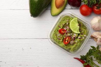 Bowl of delicious guacamole with onion, lime, chili pepper and ingredients on white wooden table, flat lay. Space for text