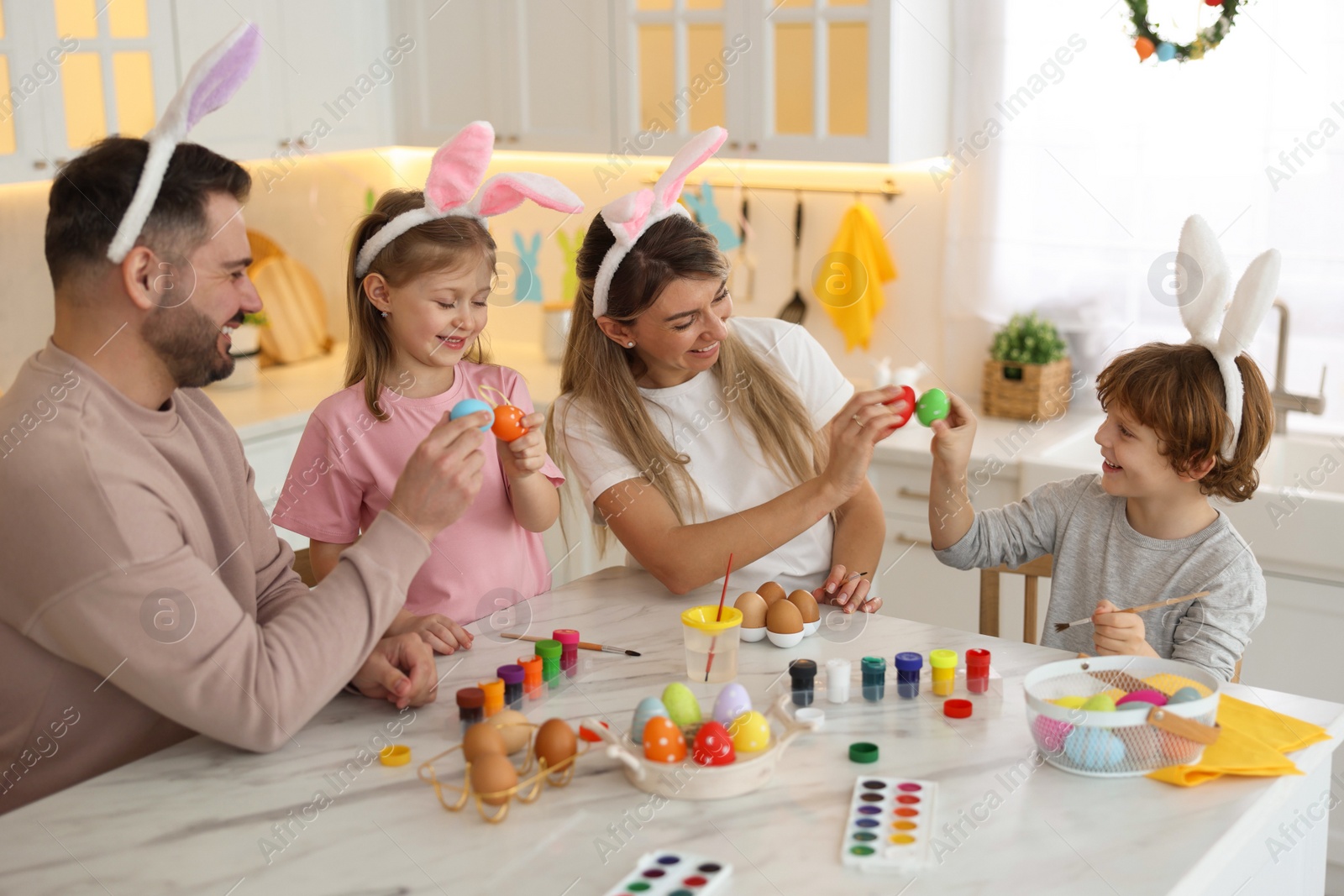 Photo of Easter celebration. Happy family with bunny ears having fun while painting eggs at white marble table in kitchen