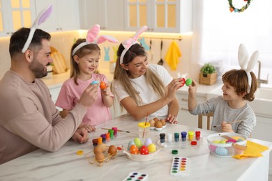 Photo of Easter celebration. Happy family with bunny ears having fun while painting eggs at white marble table in kitchen