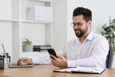 Photo of Handsome young man using smartphone while working with laptop at wooden table in office