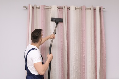 Male worker removing dust from curtains with professional vacuum cleaner indoors