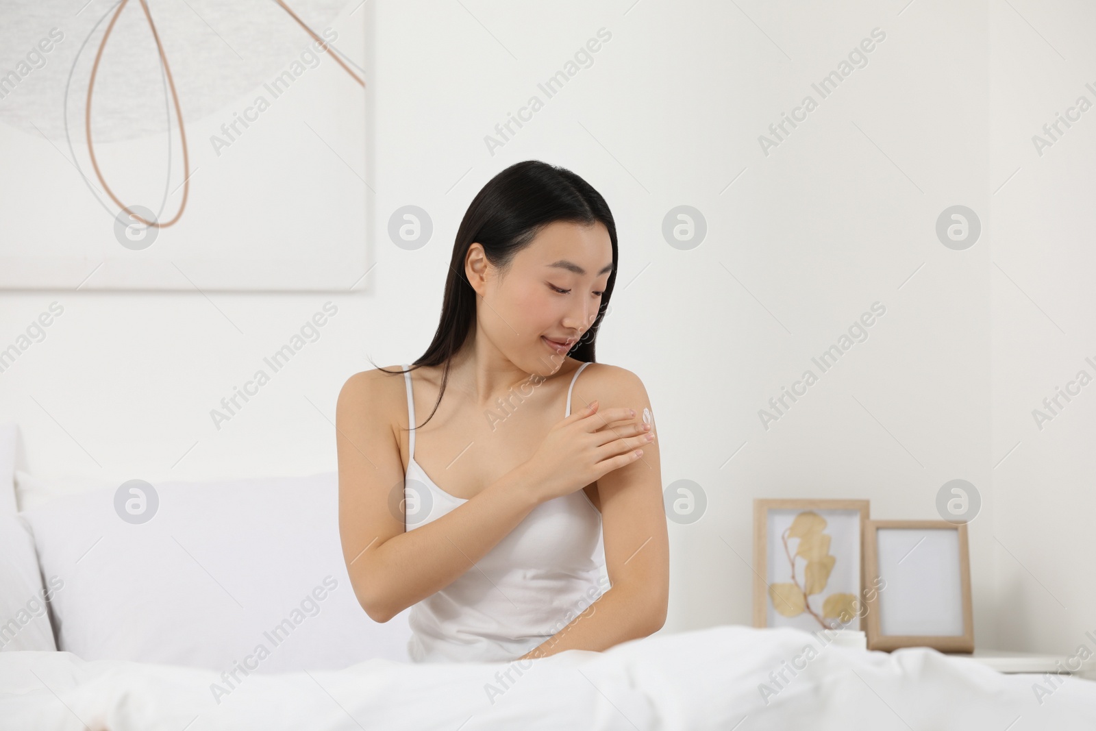 Photo of Beautiful young Asian woman applying body cream on shoulder in bedroom