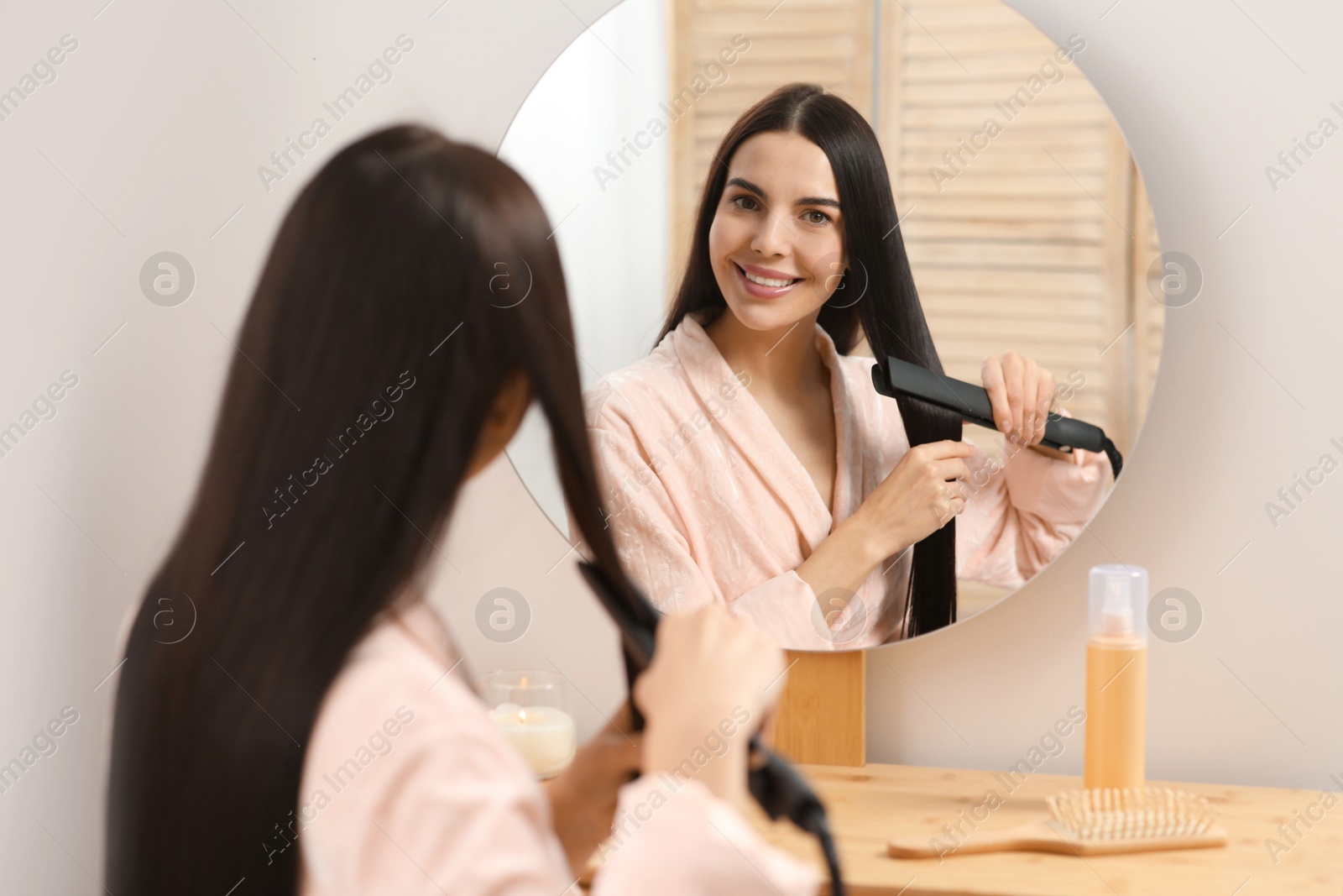 Photo of Beautiful happy woman using hair iron near mirror in room