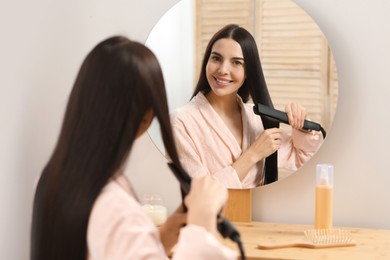 Beautiful happy woman using hair iron near mirror in room