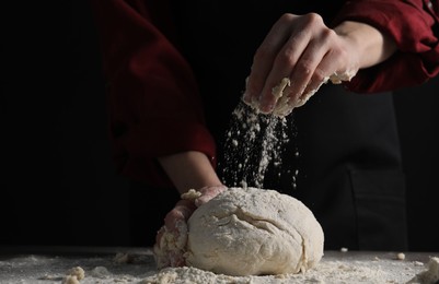 Photo of Making bread. Woman sprinkling flour over dough at table on dark background, closeup