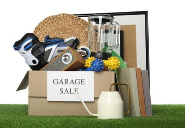 Photo of Box with sign Garage Sale and different stuff on green grass against white background