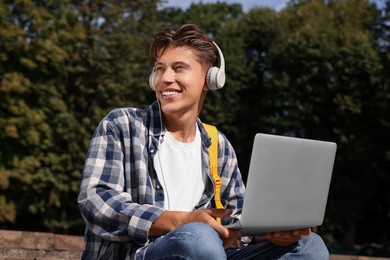 Photo of Happy young student with headphones studying with laptop on steps in park