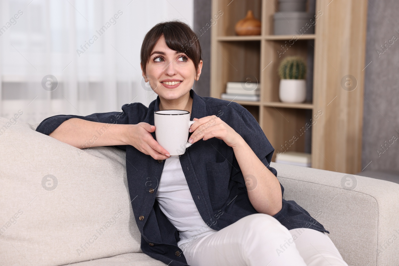 Photo of Beautiful young housewife with cup of drink on sofa at home