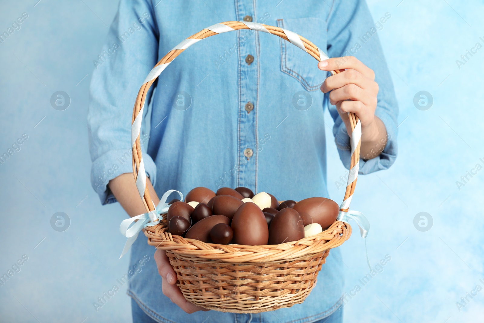 Photo of Woman holding wicker basket with chocolate Easter eggs on color background, closeup