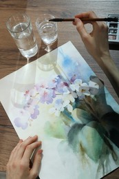 Photo of Woman painting flowers with watercolor at wooden table, above view. Creative artwork