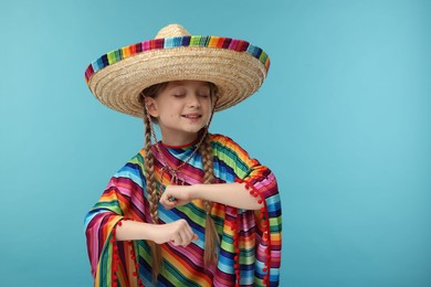 Photo of Cute girl in Mexican sombrero hat and poncho dancing on light blue background