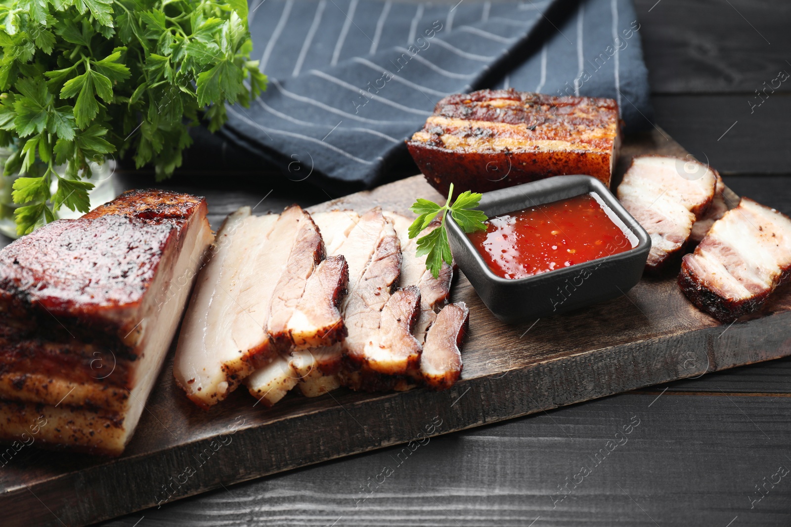 Photo of Pieces of baked pork belly served with sauce and parsley on black wooden table, closeup