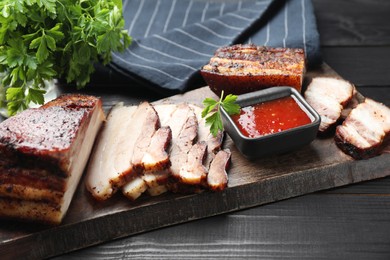 Photo of Pieces of baked pork belly served with sauce and parsley on black wooden table, closeup