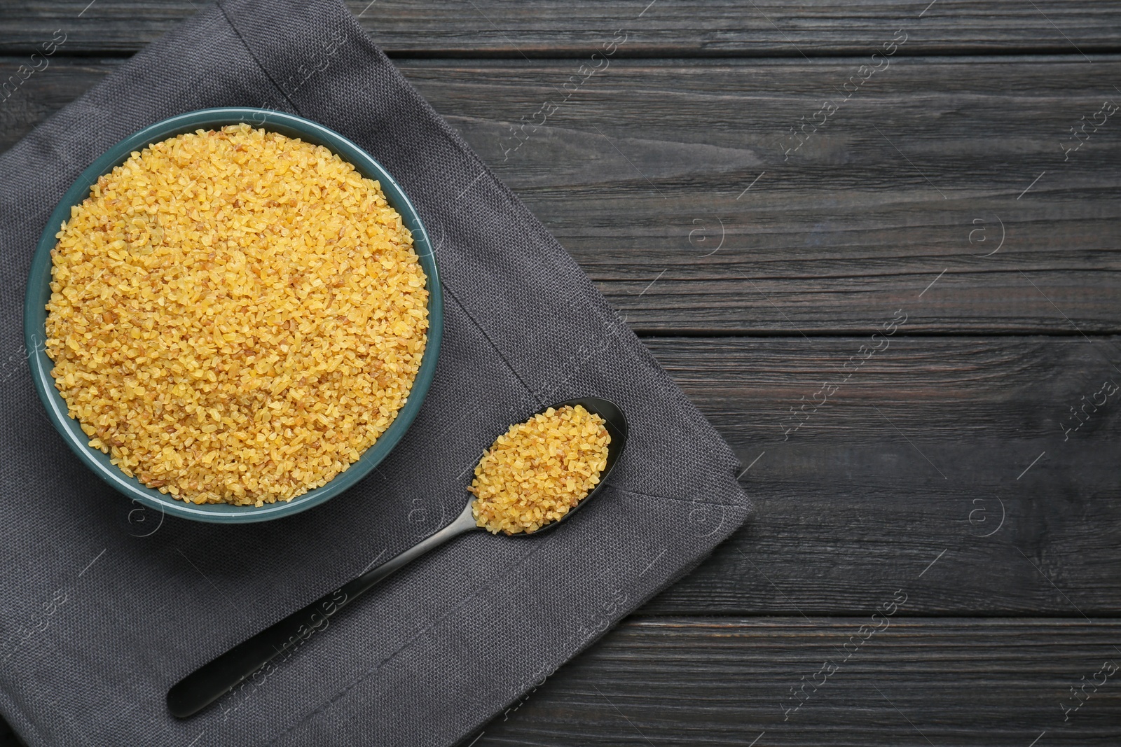 Photo of Bowl and spoon with uncooked bulgur on black wooden table, flat lay. Space for text