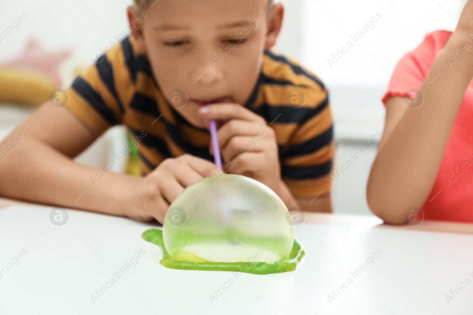 Photo of Little boy blowing slime bubble on white table indoors, closeup