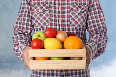 Photo of Man holding wooden crate filled with fresh vegetables and fruits against color background, closeup