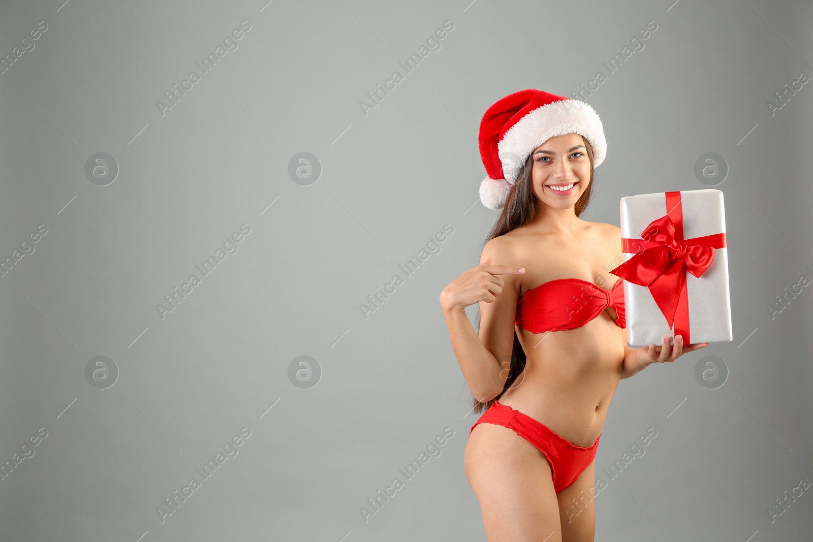 Photo of Young beautiful woman in Santa hat and swimsuit with gift box on grey background. Christmas celebration