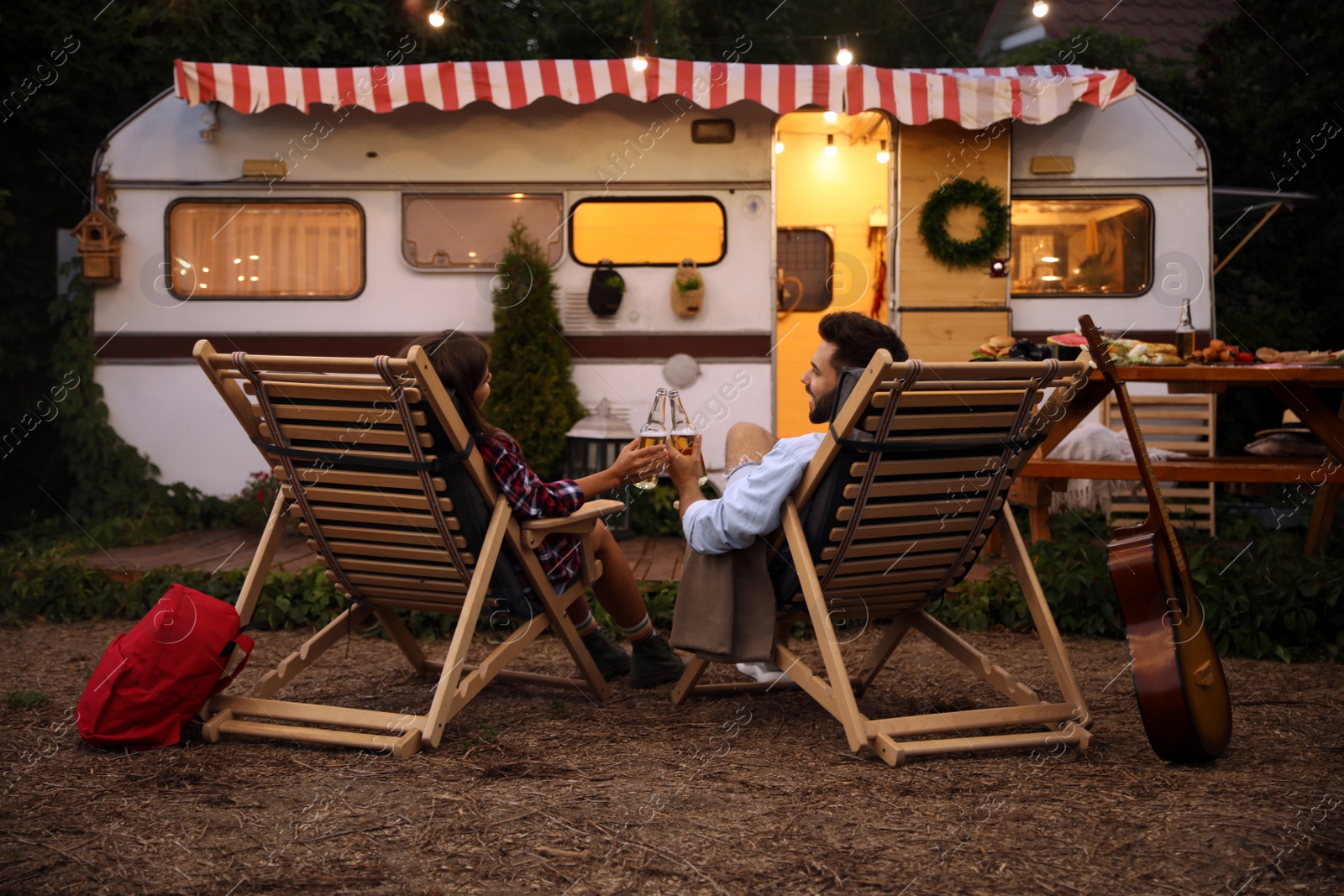 Photo of Young couple toasting with bottles of beer near trailer. Camping season