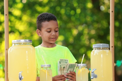 Photo of Cute little African-American boy with money at lemonade stand in park. Summer refreshing natural drink