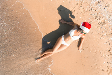 Image of Young woman wearing Santa hat and bikini on sunny beach, top view. Christmas vacation