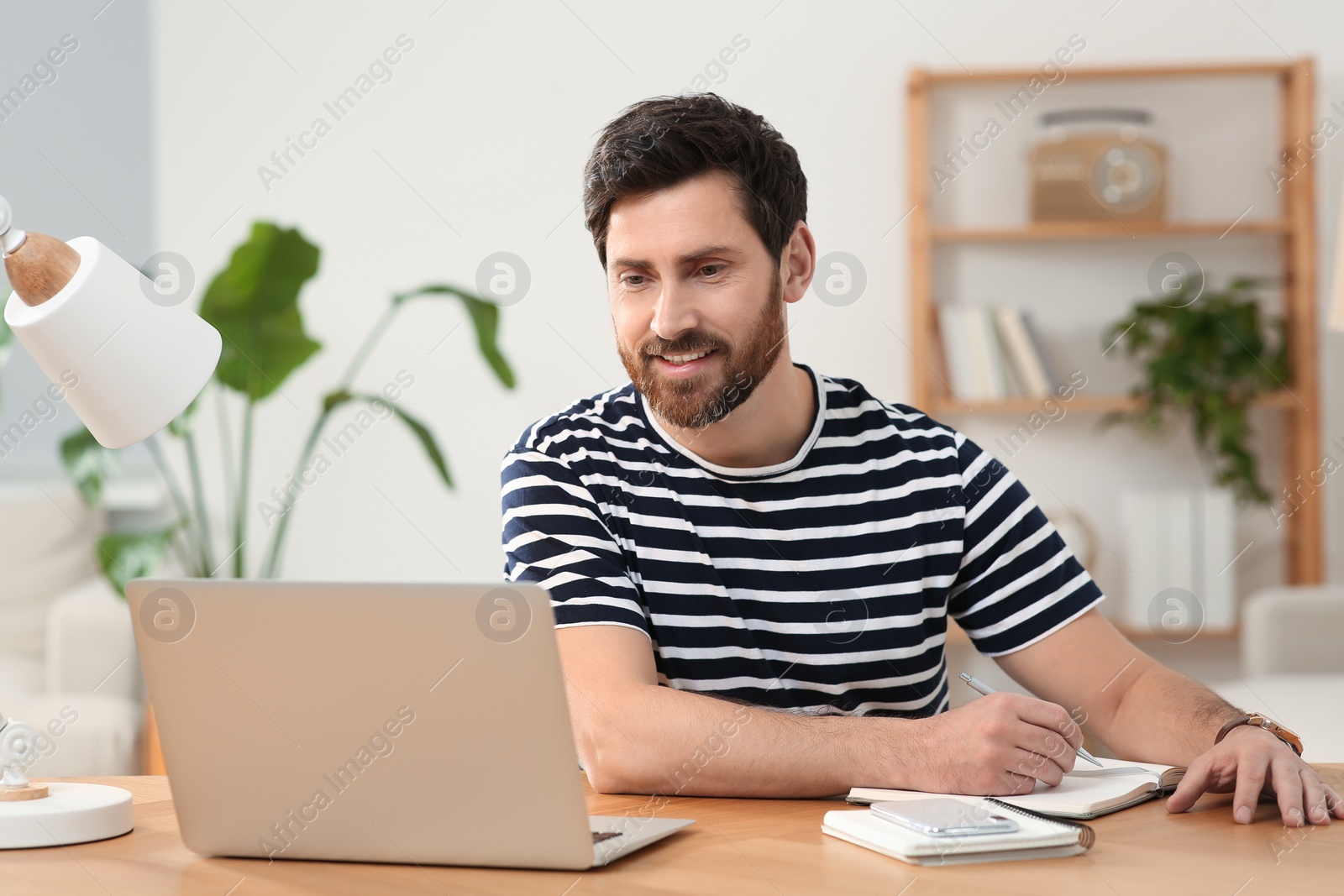 Photo of Home workplace. Happy man taking notes while working with laptop at wooden desk in room