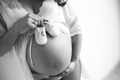 Photo of Young pregnant woman in lace nightgown holding baby shoes on light background, closeup with space for text. Black and white effect