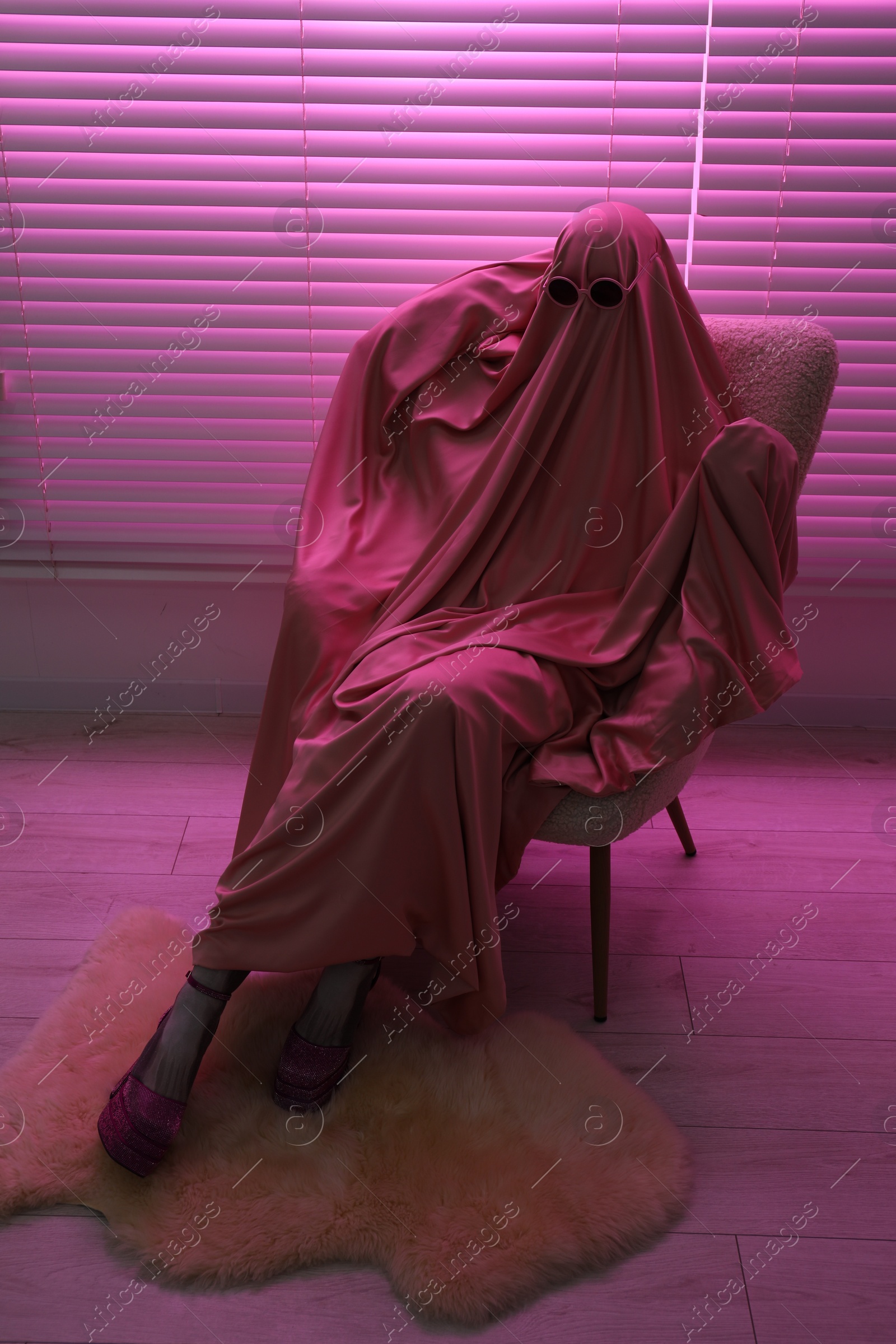 Photo of Glamorous ghost. Woman in sheet and high heel shoes on armchair in pink light