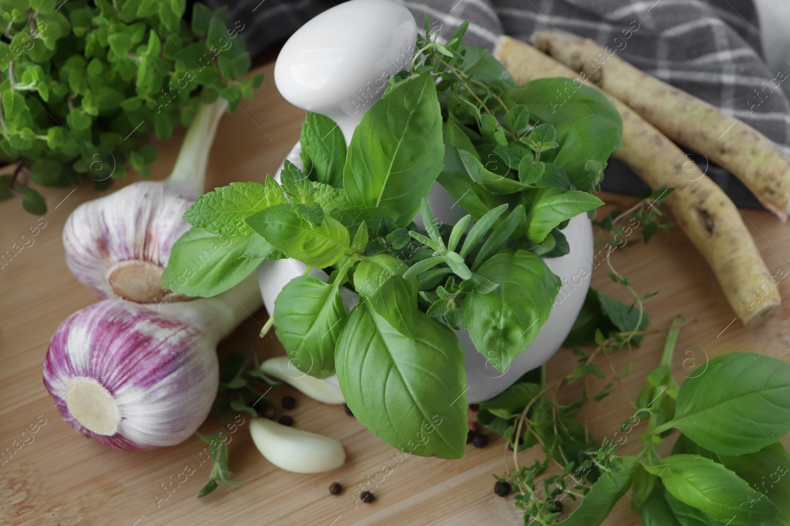 Photo of Mortar with different fresh herbs near garlic, horseradish roots and black peppercorns on wooden table, closeup