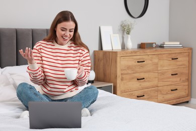 Photo of Happy woman with cup of drink and laptop on bed in bedroom