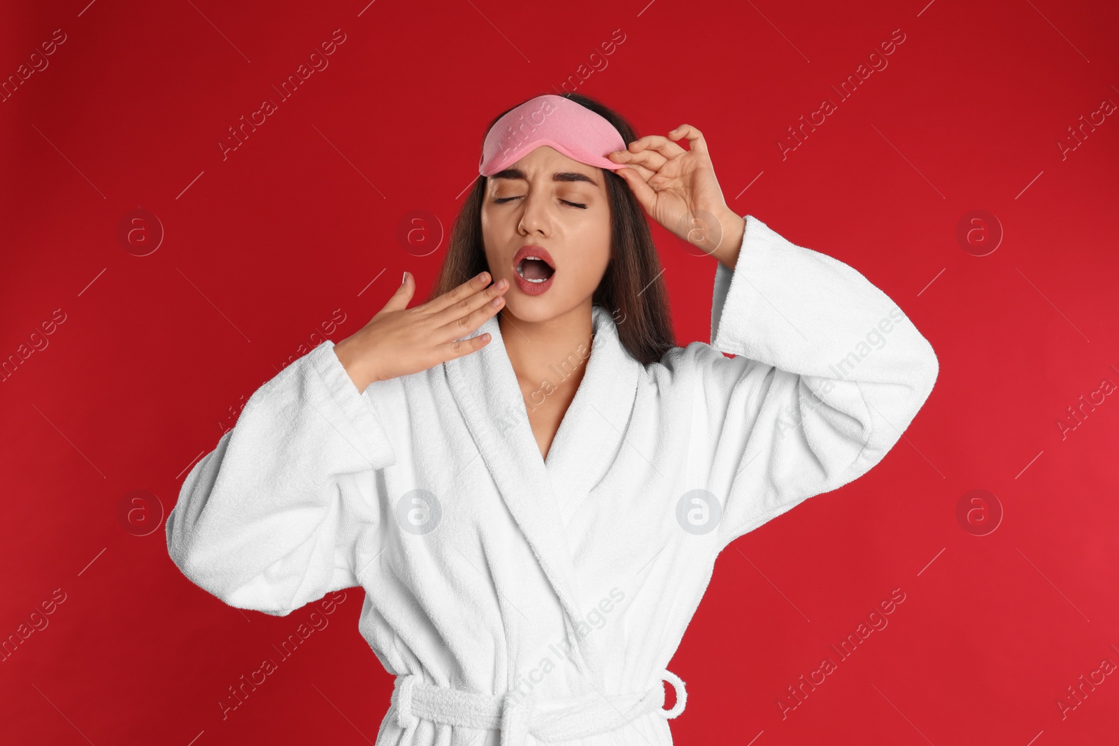 Photo of Young woman in bathrobe and eye sleeping mask yawning on red background