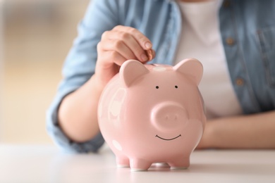 Woman putting coin into piggy bank at table indoors, closeup