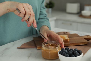Woman taking tasty nut butter onto knife at marble table, closeup