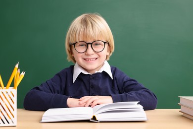 Photo of Happy little school child sitting at desk with book near chalkboard
