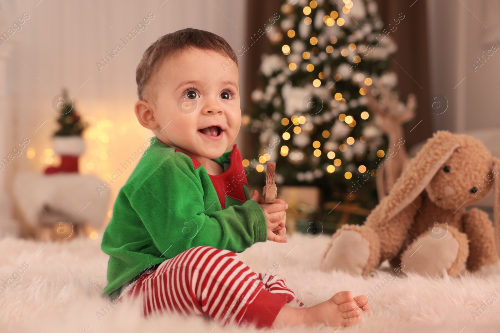 Photo of Baby wearing cute elf costume on floor in room decorated for Christmas