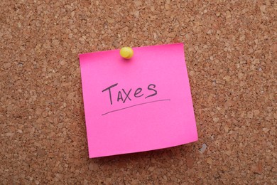 Photo of Pink paper note with phrase Taxes pinned to cork board, closeup