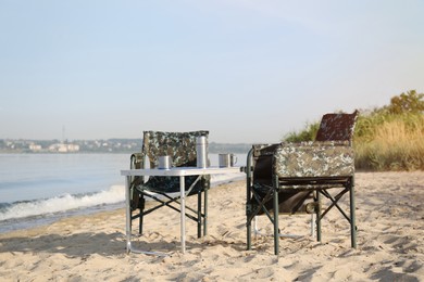 Photo of Camouflage fishing chairs and table with metal cups on sandy beach near river