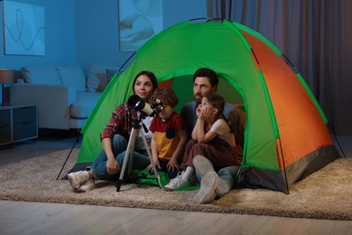 Photo of Happy family using telescope to look at stars while sitting in camping tent indoors