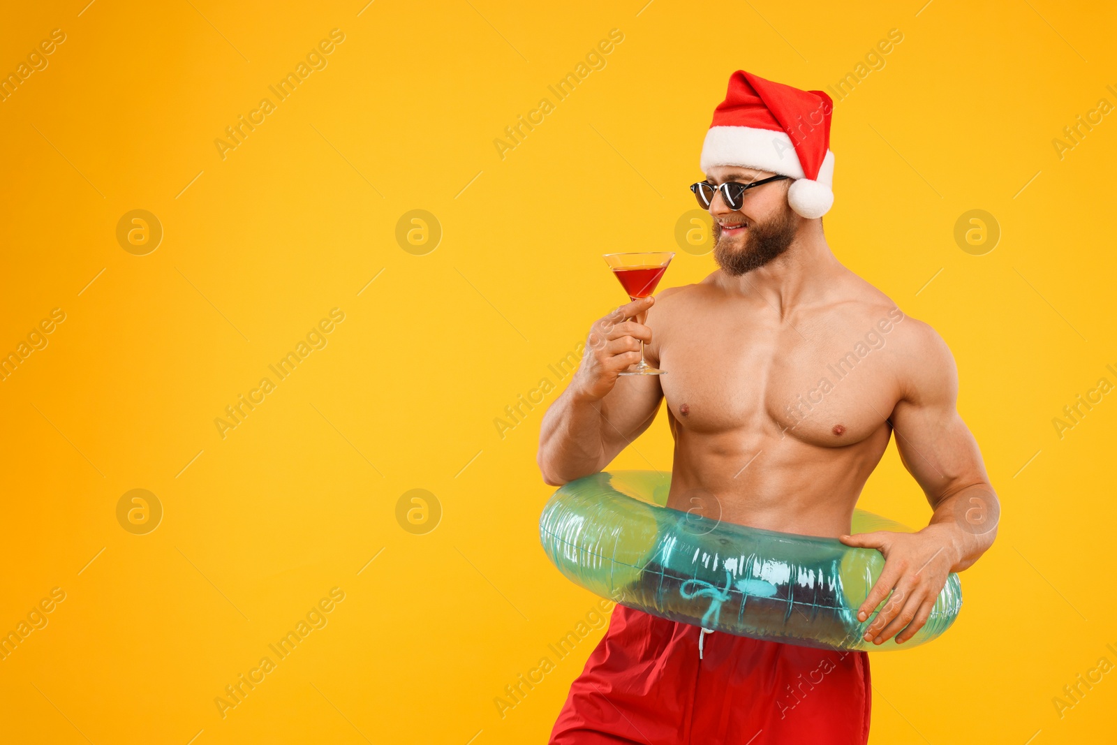 Photo of Muscular young man in Santa hat holding inflatable ring and cocktail on orange background, space for text