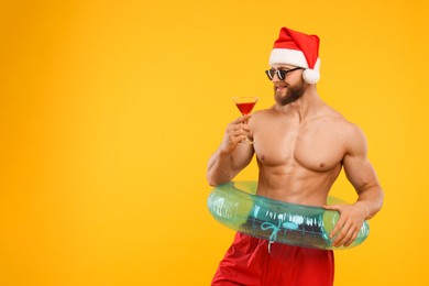 Muscular young man in Santa hat holding inflatable ring and cocktail on orange background, space for text