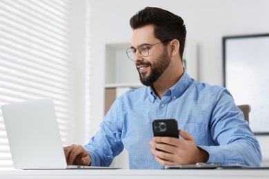 Handsome young man using smartphone while working with laptop at white table in office