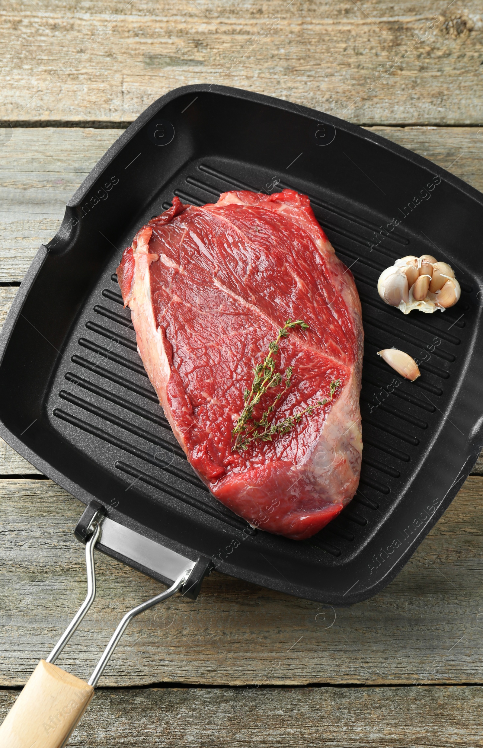 Photo of Grill pan with piece of raw beef meat, garlic and thyme on wooden table, top view