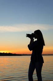 Photo of Young female photographer taking photo of riverside sunset with professional camera outdoors