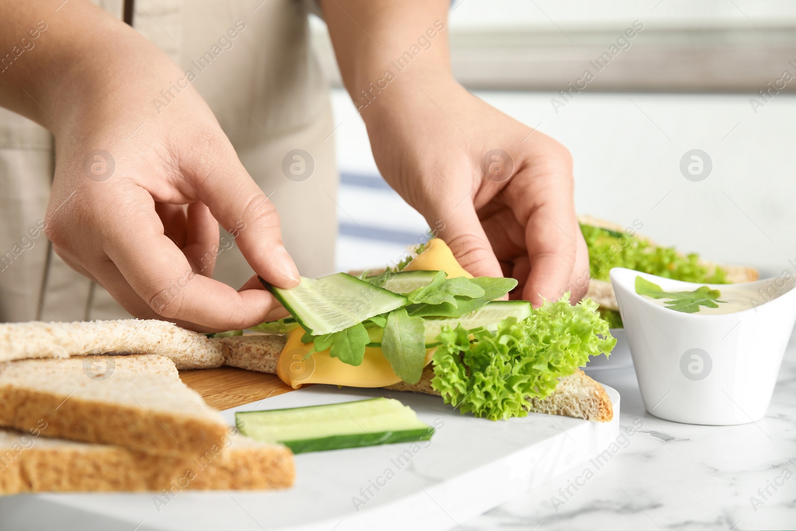 Photo of Woman adding cucumber to tasty sandwich at white marble table, closeup