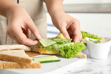 Photo of Woman adding cucumber to tasty sandwich at white marble table, closeup