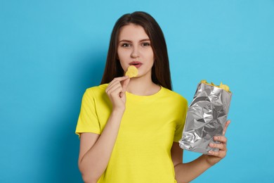 Pretty young woman eating tasty potato chips on light blue background