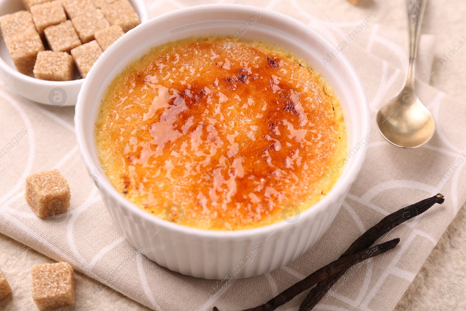 Photo of Delicious creme brulee in bowl, vanilla pods and sugar cubes on table, closeup