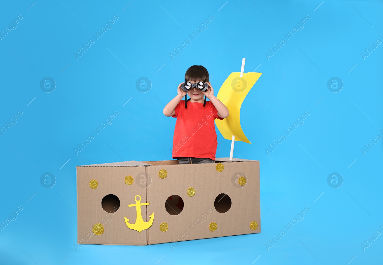 Photo of Cute little child playing with binoculars and cardboard ship on light blue background