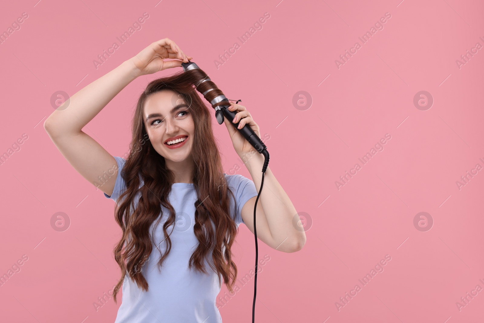 Photo of Beautiful young woman using curling hair iron on pink background, space for text