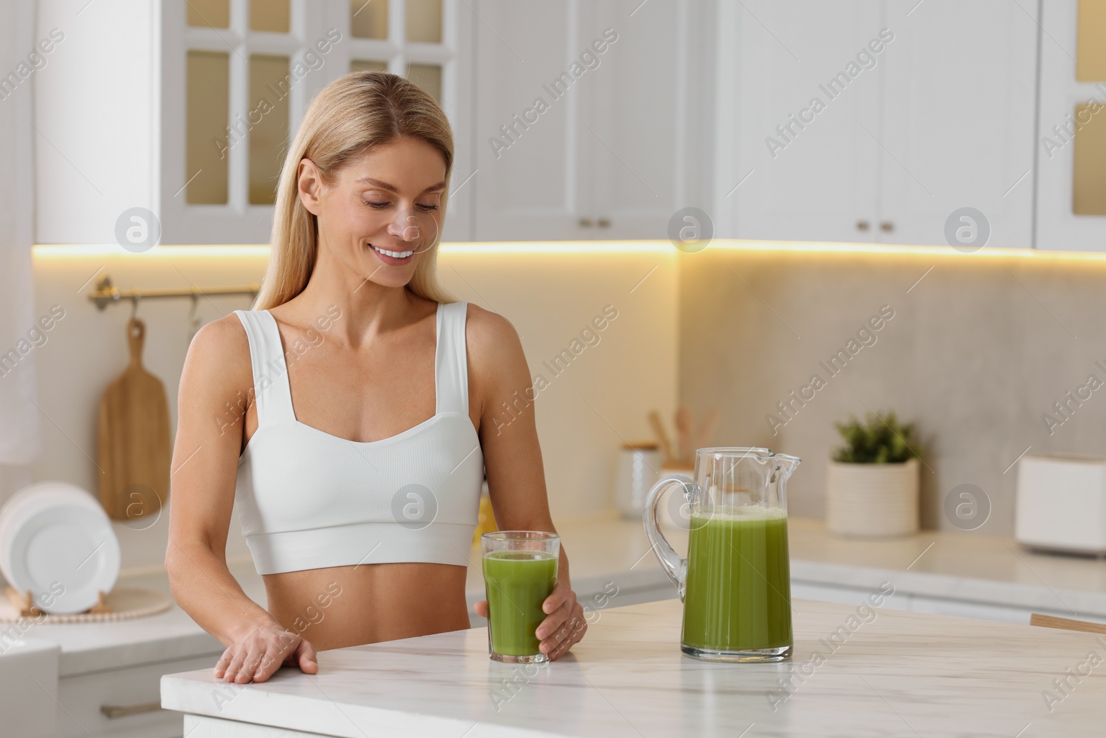 Photo of Happy woman with glass of fresh celery juice at table in kitchen